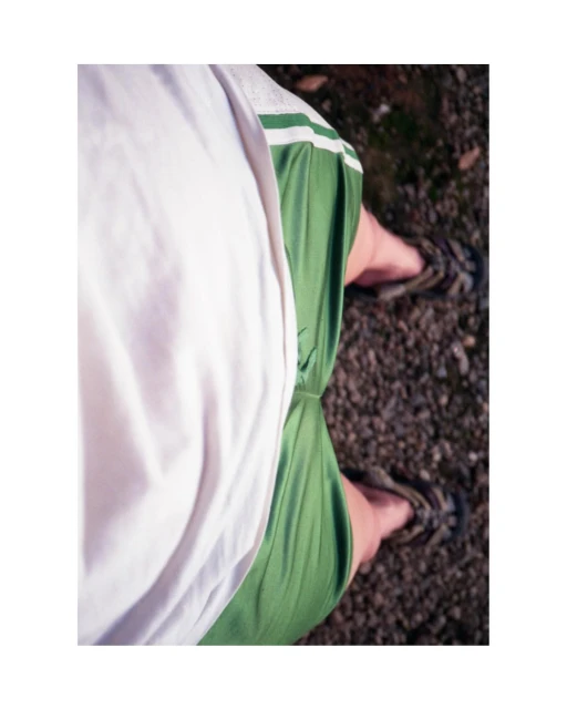 Overhead shot of a person wearing a white shirt, green shorts, and sandals standing on a gravel surface, capturing casual style.