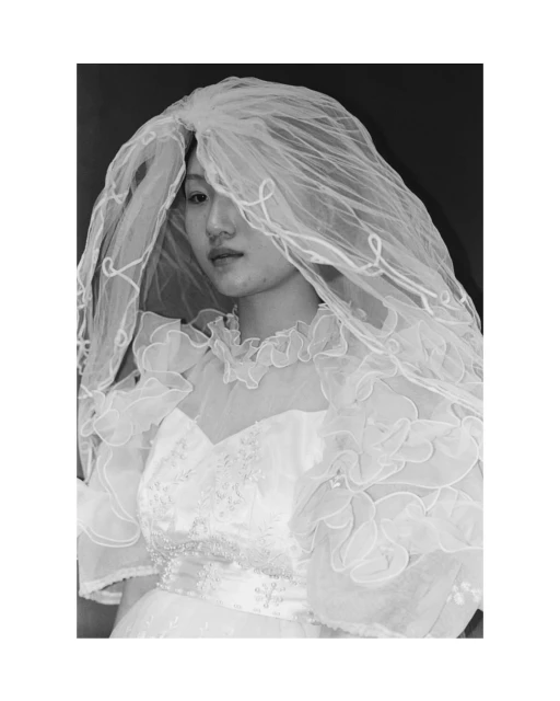 Black and white portrait of a young woman in a white wedding dress and veil against a dark background, with a soft, romantic aesthetic.