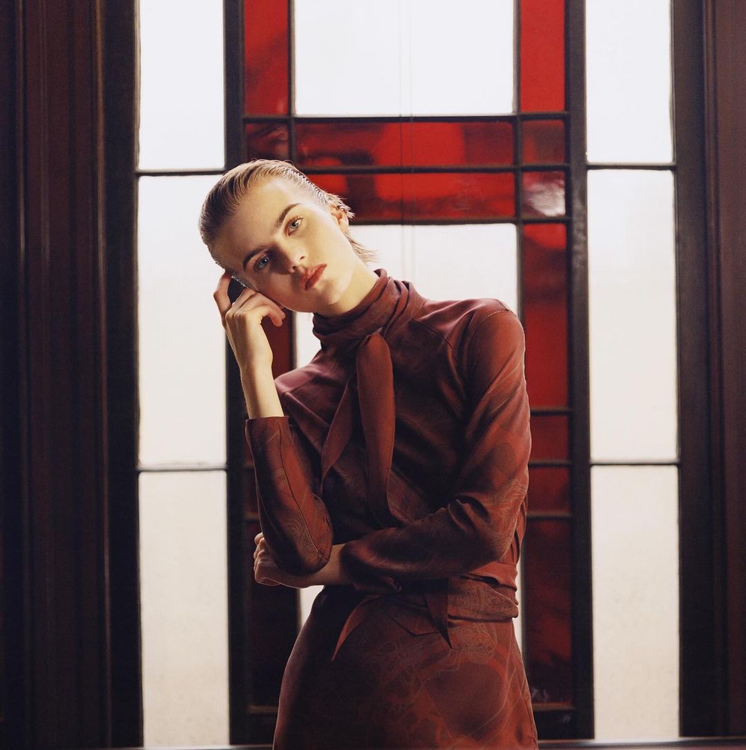 Woman in burgundy dress posing against a window with red glass panes. Elegant fashion shot with natural lighting and soft, muted tones.