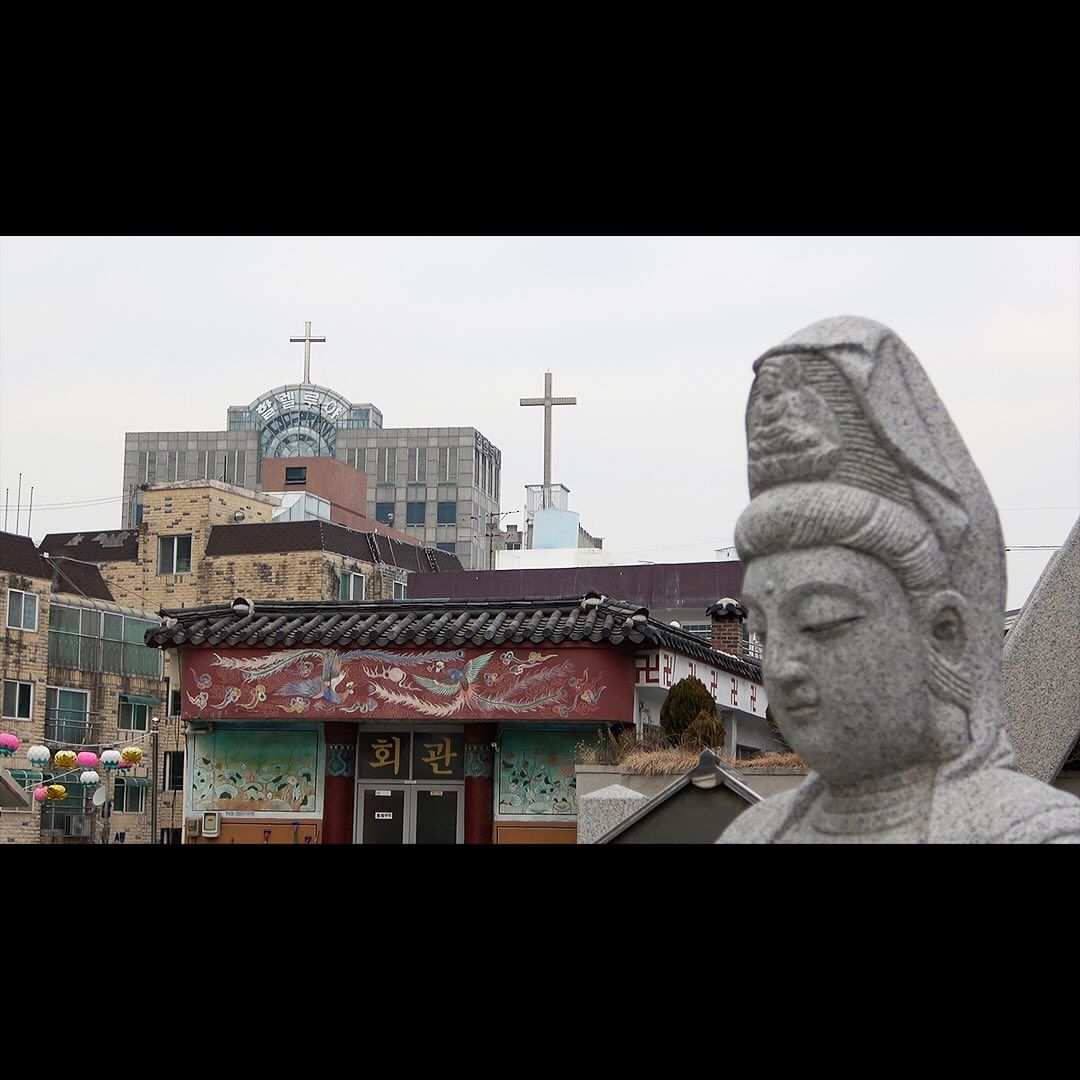 Stone statue of a head with temple and cross-topped buildings in the background, against a cloudy sky in a city setting.