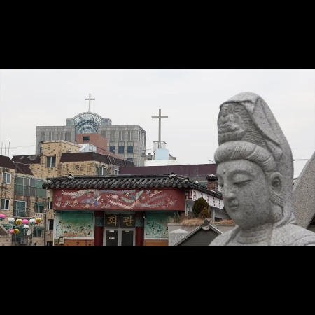 Stone statue of a head with temple and cross-topped buildings in the background, against a cloudy sky in a city setting.