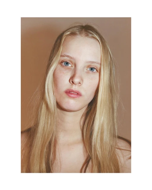 Portrait of a blonde woman with blue eyes gazing directly at the camera against a neutral peach-colored background in a studio setting.