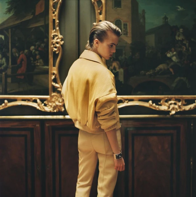 Young adult stands indoors wearing a beige jacket and pants, looking at an old painting in a decorative gold frame.