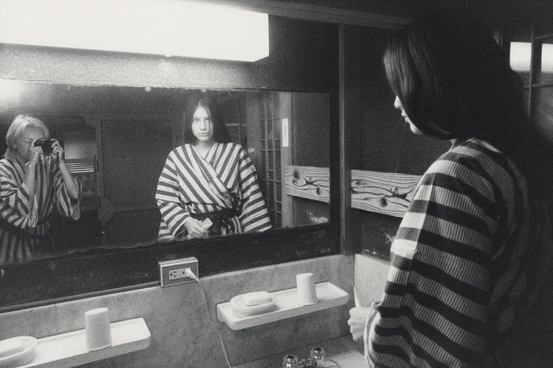 Black and white photo of a woman in a striped robe looking into a mirror with her reflection and another person taking her picture.