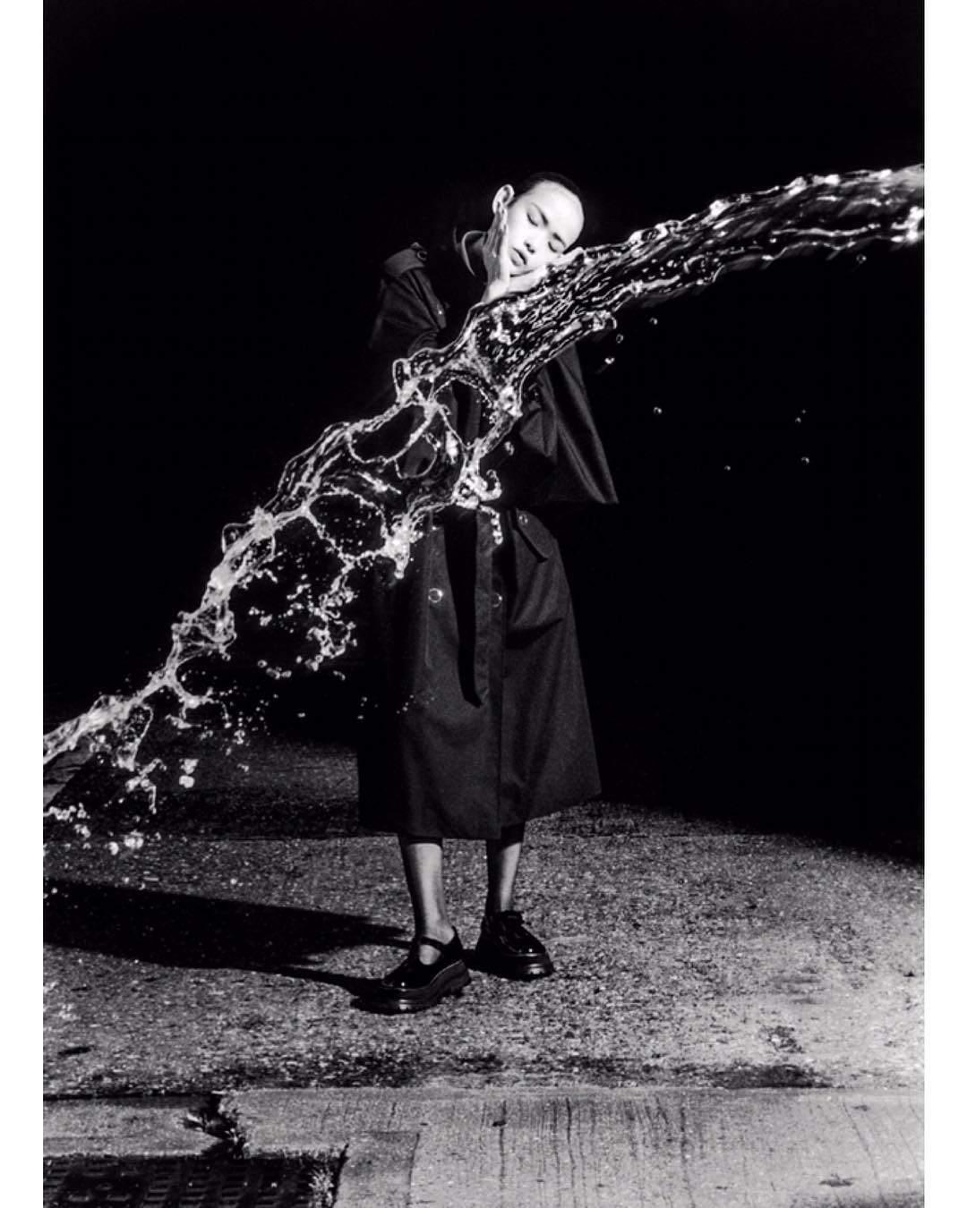 Monochrome fashion shot of a bald adult model with eyes closed as water cascades along their face and body in a black coat.