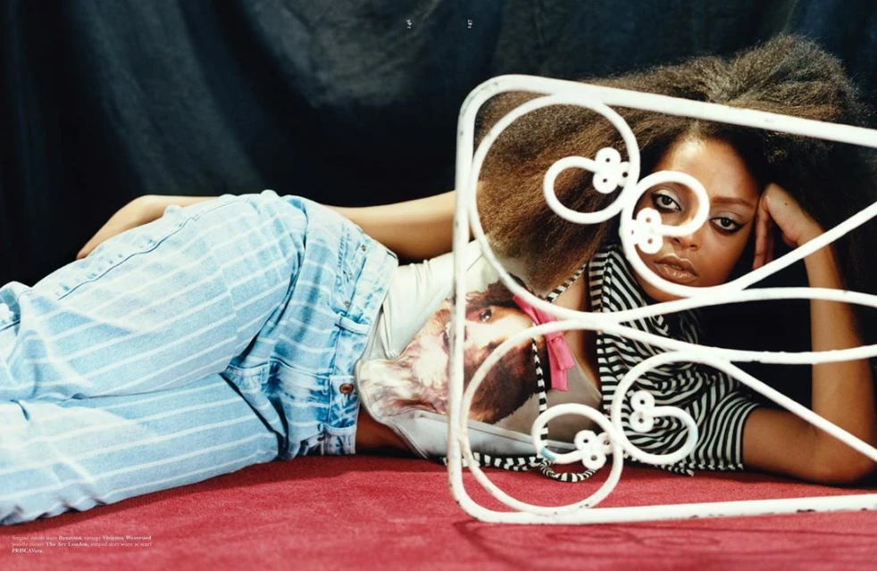 A woman with big curly hair, wearing a striped top and striped jeans, is reclining next to a wrought-iron headboard on a red surface.