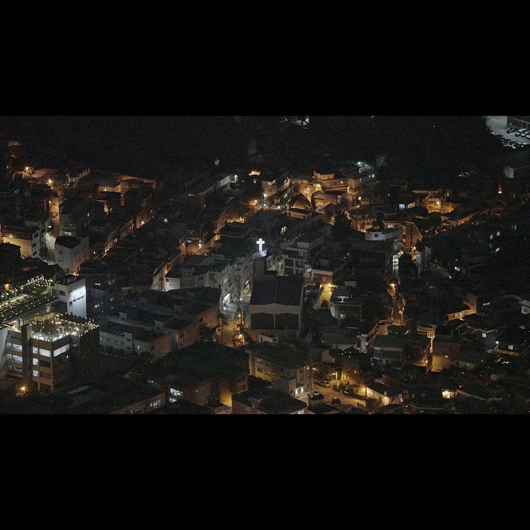 Night aerial view of a European town, featuring dark buildings illuminated by streetlights and a church with a bright cross.