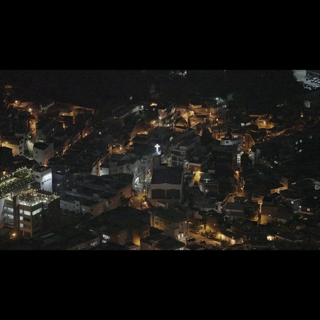 Night aerial view of a European town, featuring dark buildings illuminated by streetlights and a church with a bright cross.