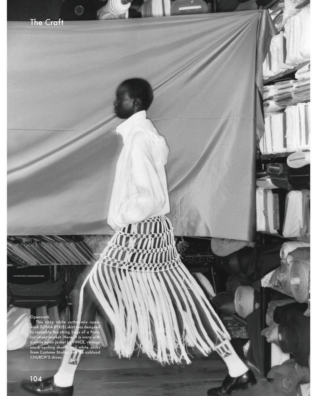 Fashion editorial shot in black and white, featuring a Black model in a unique skirt with long fringes, against a draped backdrop.