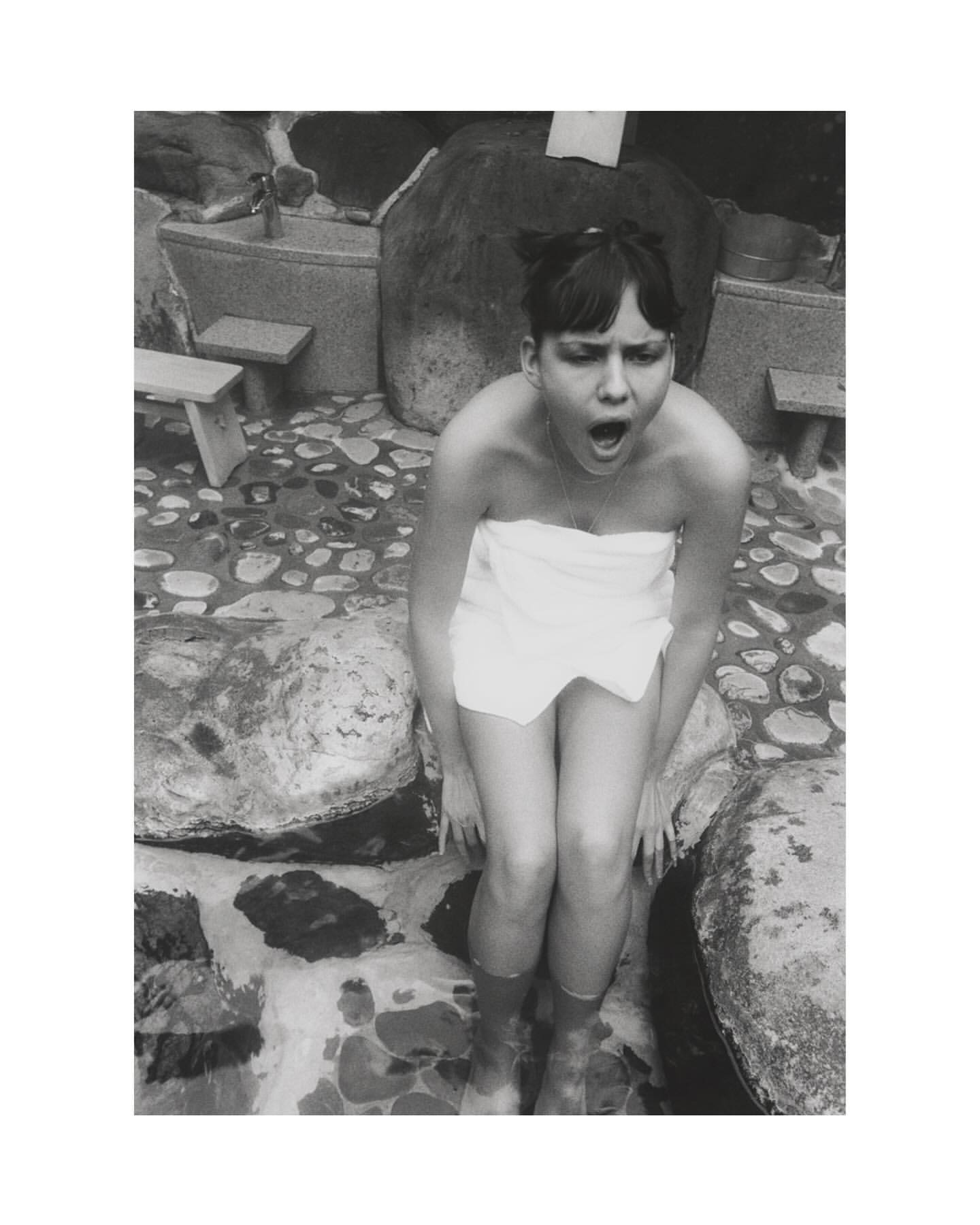 Black and white portrait of a surprised young woman wearing a towel, seated outside near stones, with her feet in water.