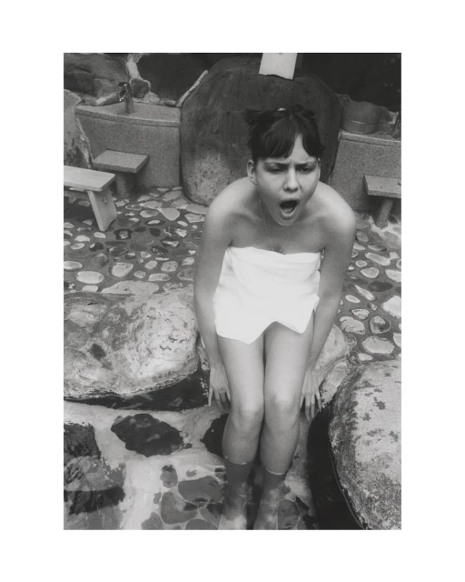 Black and white portrait of a surprised young woman wearing a towel, seated outside near stones, with her feet in water.