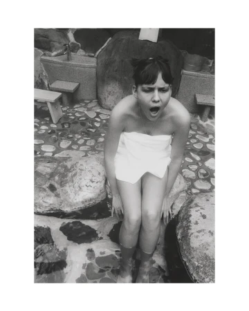 Black and white portrait of a surprised young woman wearing a towel, seated outside near stones, with her feet in water.