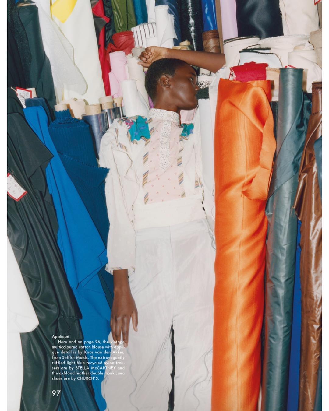 Fashion model poses in a white outfit among colorful rolls of textile fabrics in a photo studio with bright, diffused lighting.