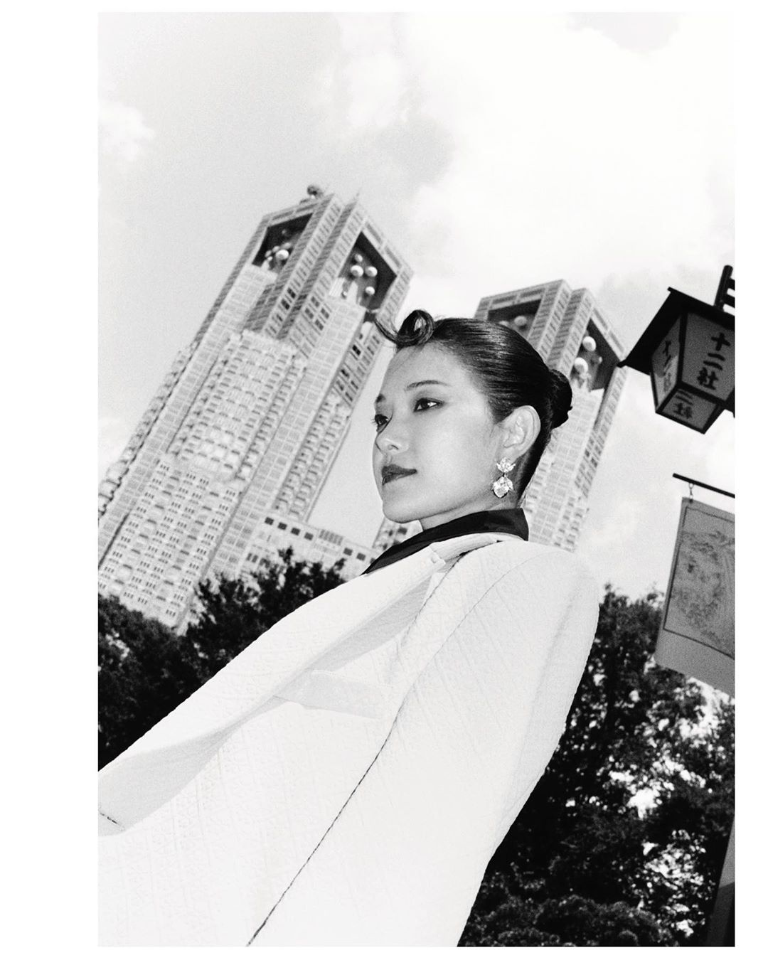 Black and white portrait of an Asian woman in a light-colored jacket, standing in front of modern architecture on a cloudy day.