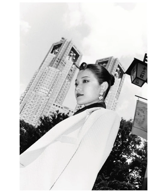 Black and white portrait of an Asian woman in a light-colored jacket, standing in front of modern architecture on a cloudy day.