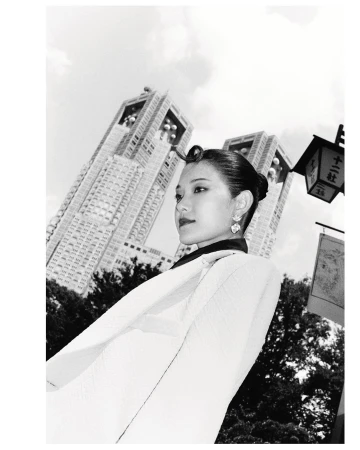 Black and white portrait of an Asian woman in a light-colored jacket, standing in front of modern architecture on a cloudy day.
