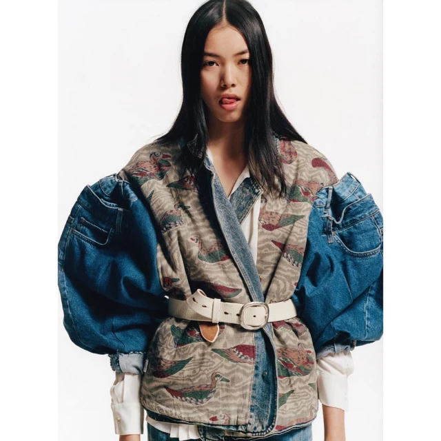 Portrait of a young Asian woman wearing a patterned and denim jacket with oversized denim sleeves, standing against a white backdrop.