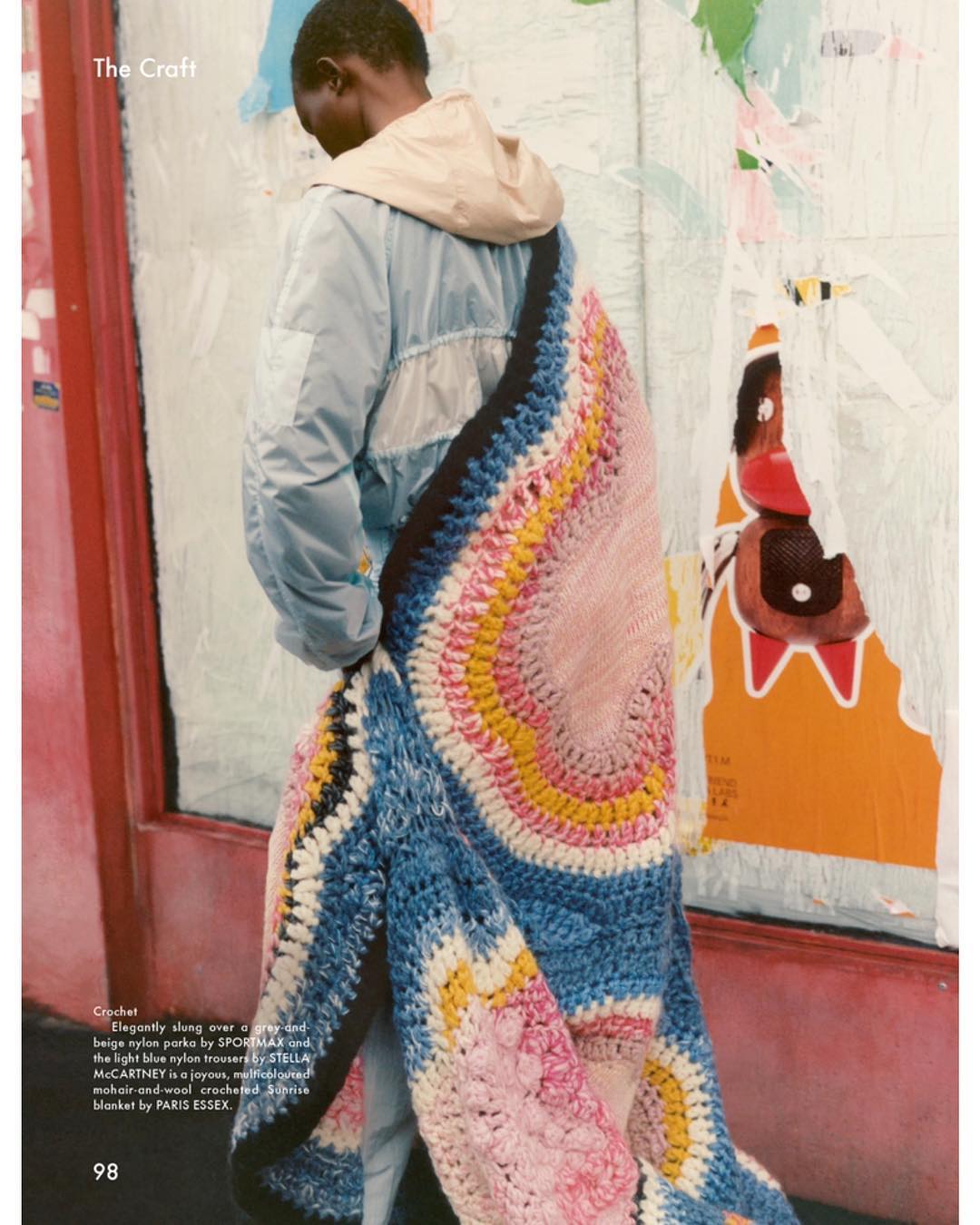 An adult male is draped in a crocheted blanket, accessorizing a light blue nylon jacket and beige hooded top, set against a textured background.