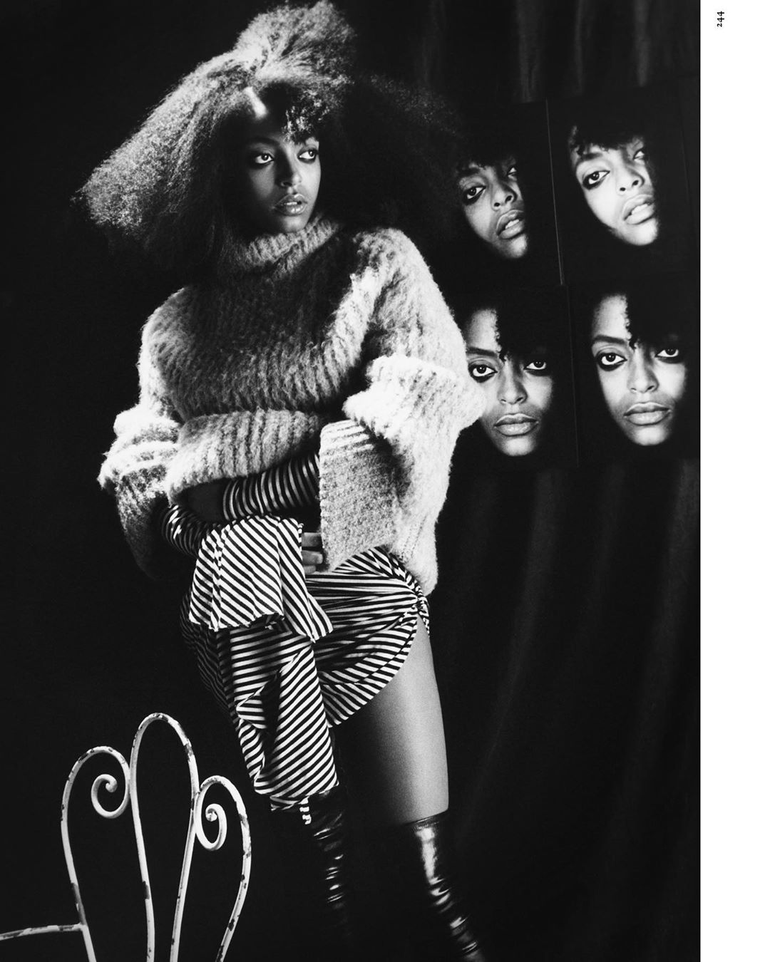 Monochrome fashion portrait of an adult woman with a voluminous afro wearing an oversized sweater, striped skirt, and shiny boots.
