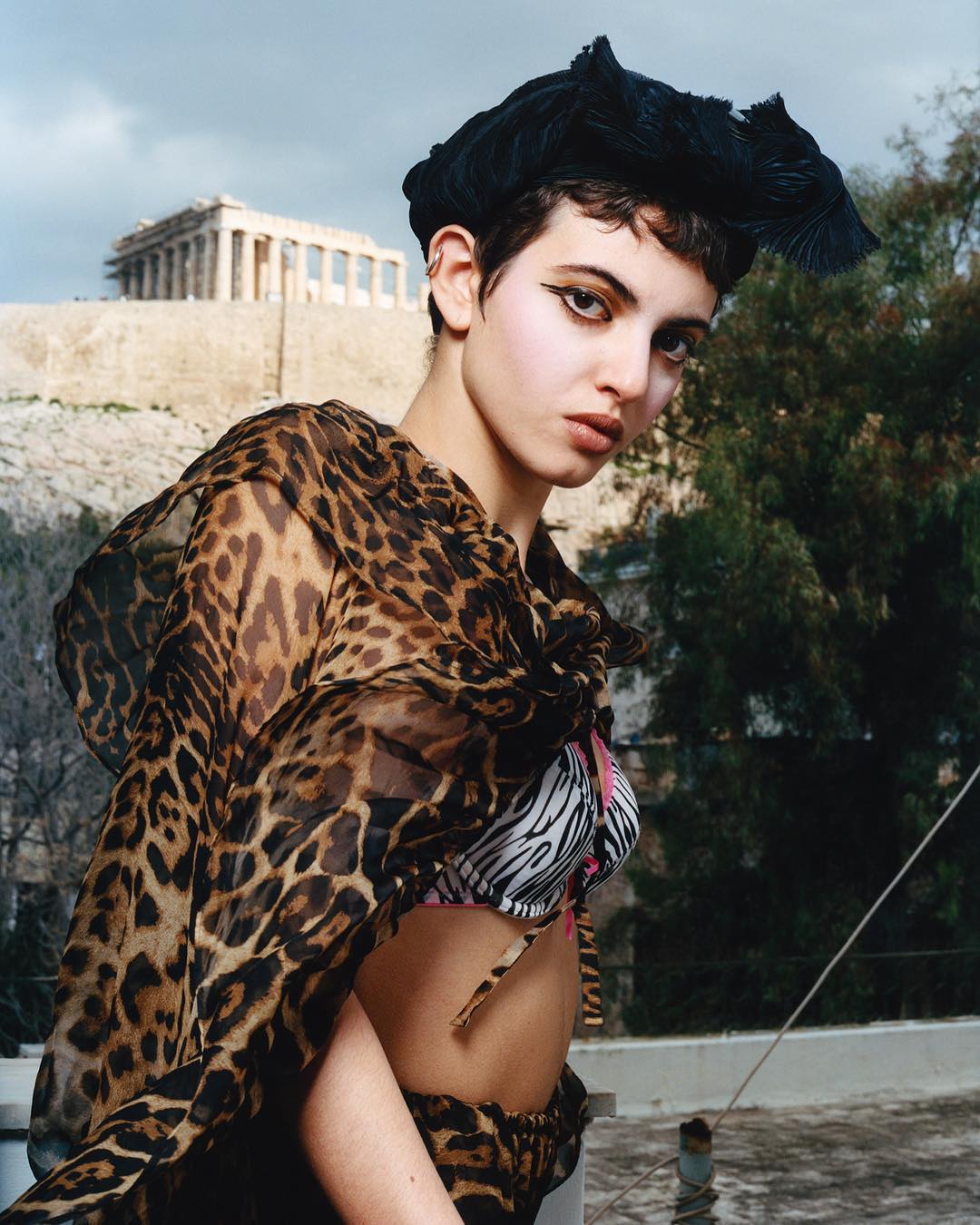 Woman in leopard-print shawl poses with Acropolis backdrop, showcasing high-fashion and architectural contrast under soft daylight.