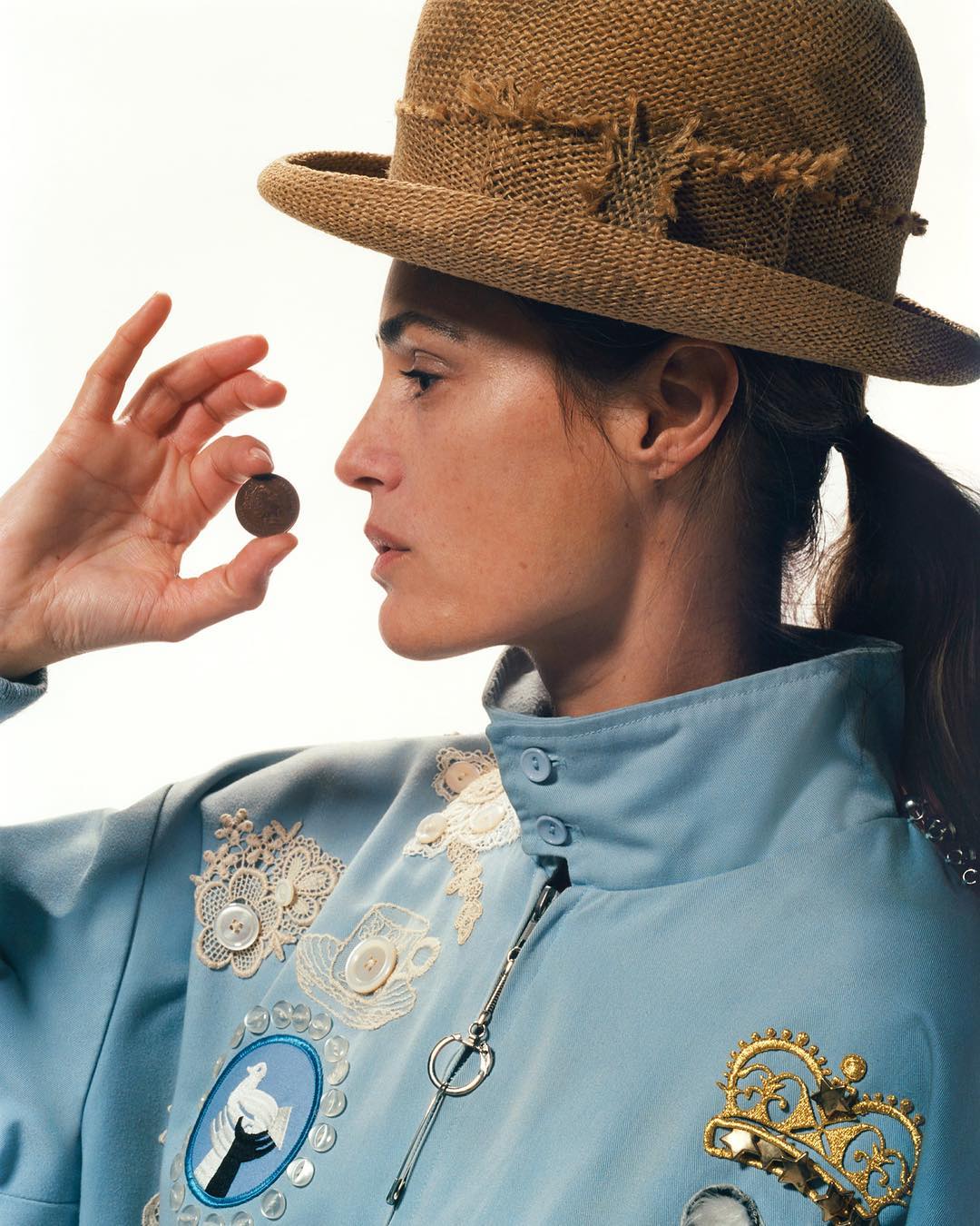 Portrait of an adult female wearing a straw hat and light blue jacket, holding a coin, posing in a photo studio with a white backdrop.