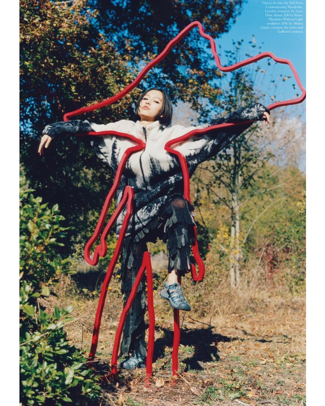 An Asian woman poses in a forest wearing a jacket and leggings with a red sculpture art installation outlining her body.