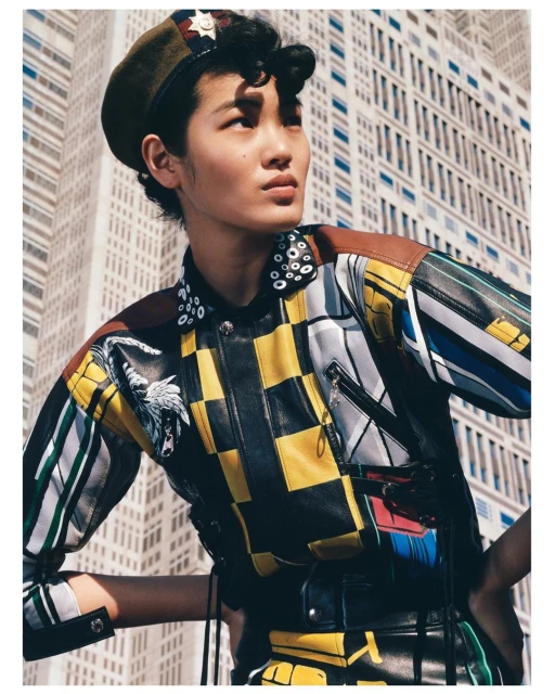 Fashionable adult with a beret, in a color-blocked leather jacket with an image of a wolf, poses in front of a high-rise building.