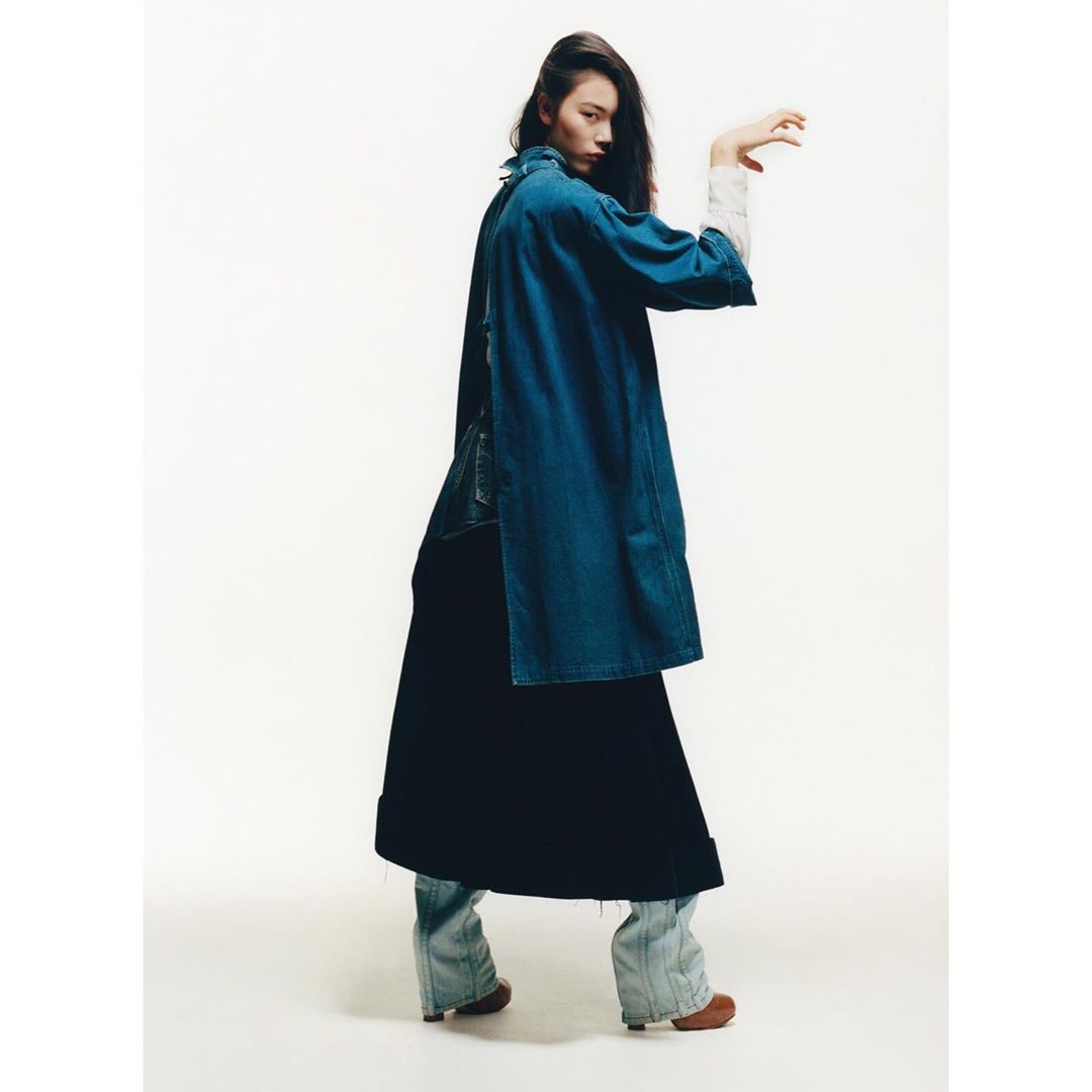 Model in blue denim coat and dark skirt posing against a white seamless background