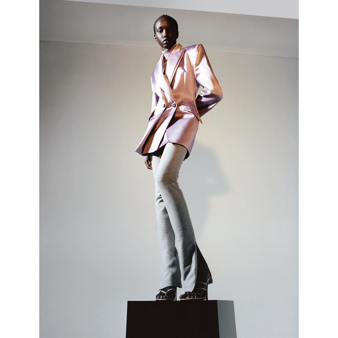 A fashion shot of an African American woman in a purple satin blazer and grey pants, standing on a dark box against a plain backdrop.