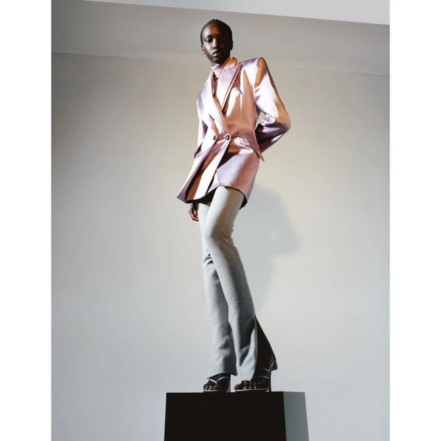 A fashion shot of an African American woman in a purple satin blazer and grey pants, standing on a dark box against a plain backdrop.