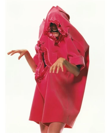 Person wearing oversized pink coat with ruffles against white background. The coat covers the head and reveals hands and part of legs.