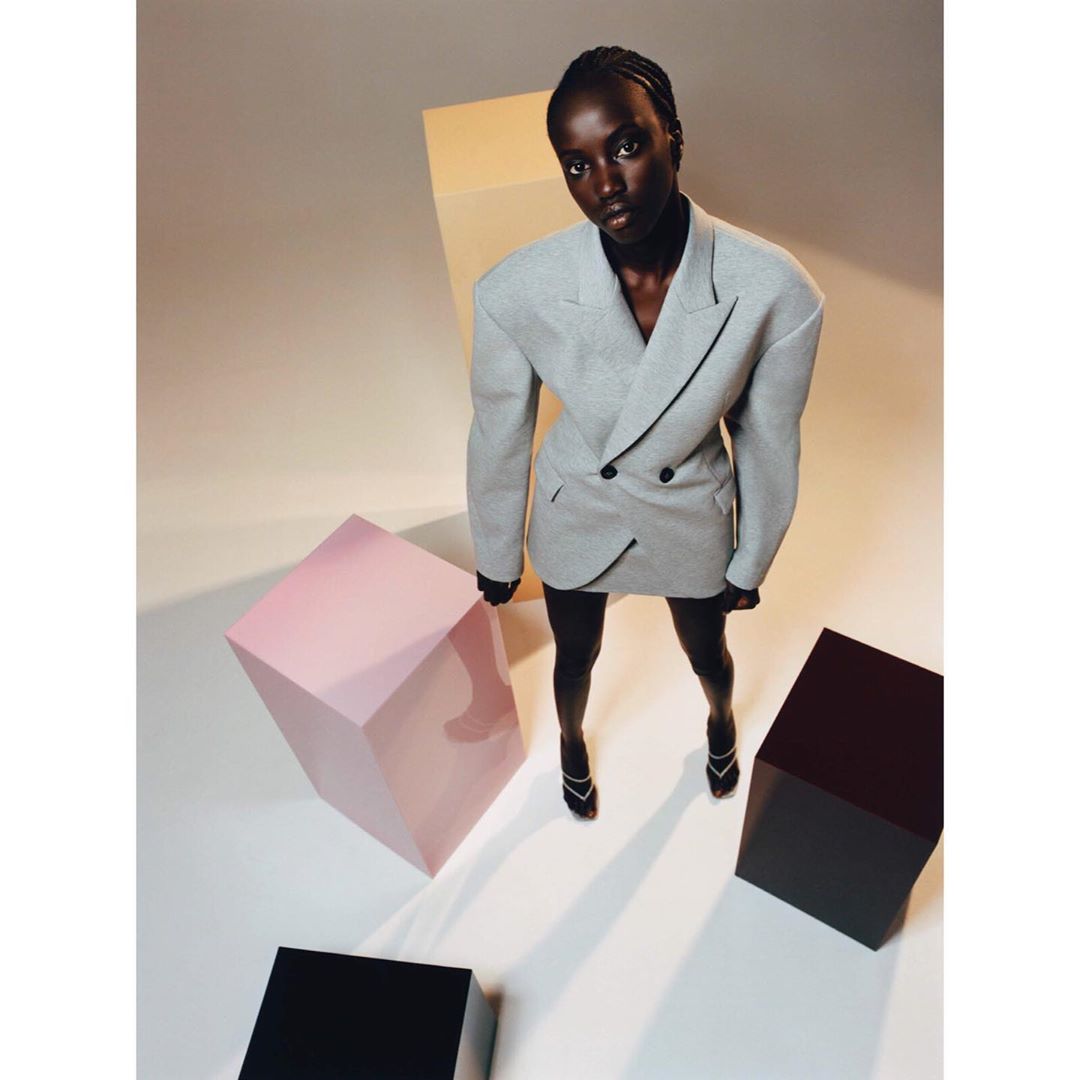 A stylish Black woman in a gray blazer dress and heels poses amid geometric blocks under studio lights, showcasing modern fashion.