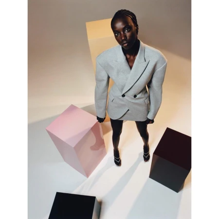 A stylish Black woman in a gray blazer dress and heels poses amid geometric blocks under studio lights, showcasing modern fashion.
