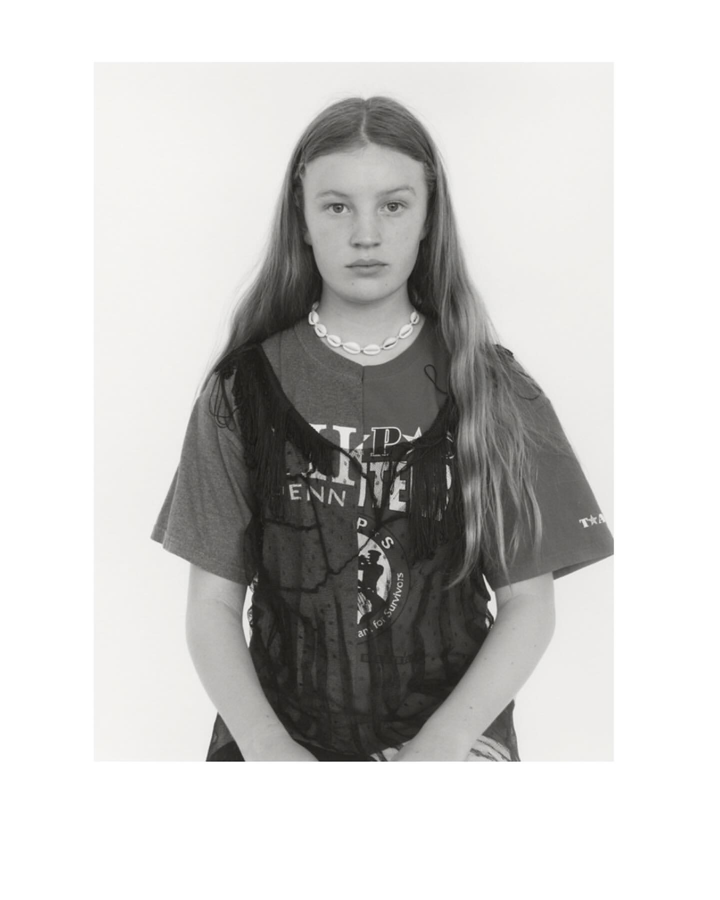 Portrait of a young woman with long hair, wearing a printed T-shirt and a shell necklace, looking directly at the camera against a white background.