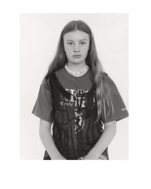 Portrait of a young woman with long hair, wearing a printed T-shirt and a shell necklace, looking directly at the camera against a white background.