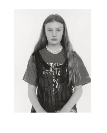 Portrait of a young woman with long hair, wearing a printed T-shirt and a shell necklace, looking directly at the camera against a white background.