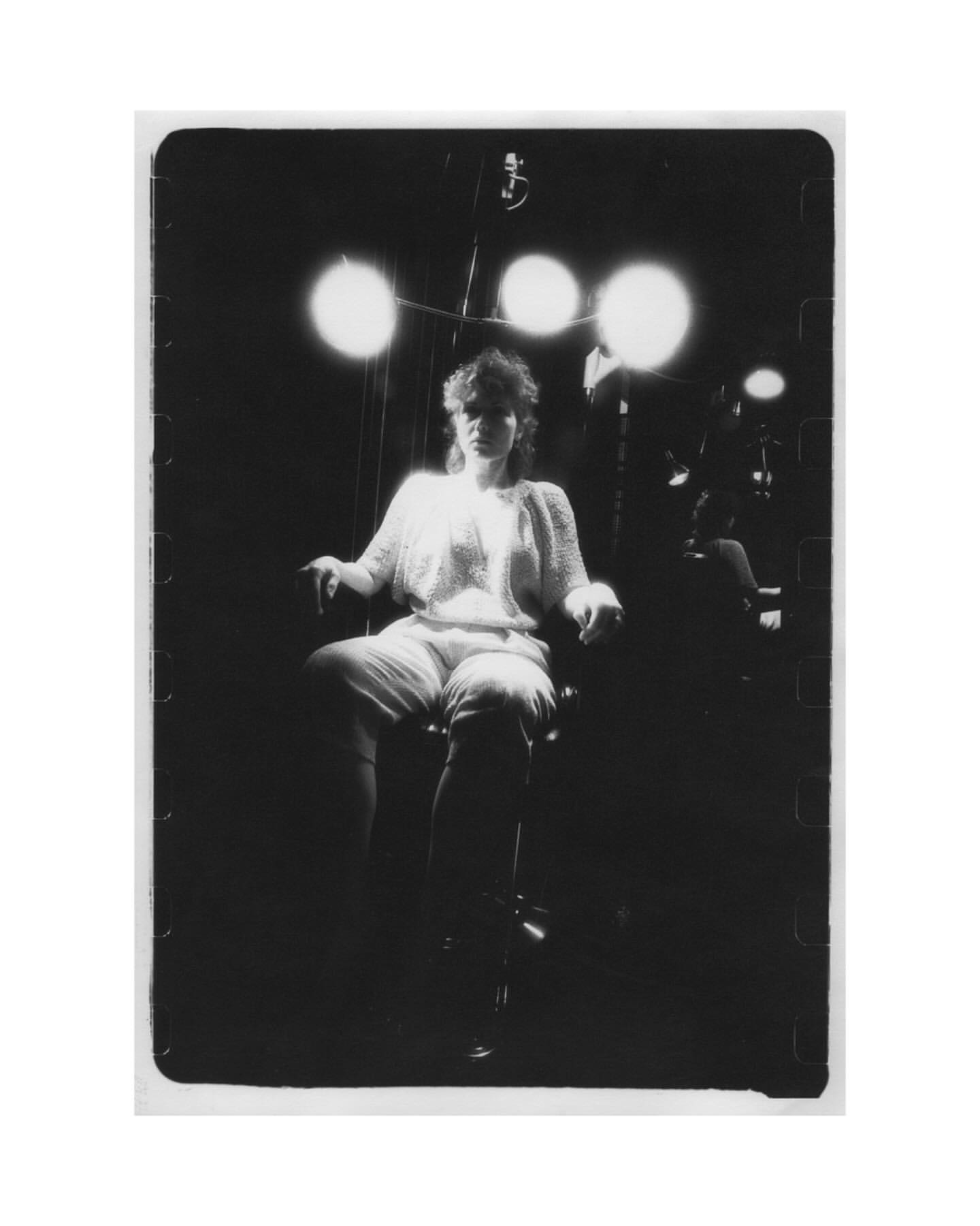 Monochrome portrait of a person sitting in a chair under bright lights, wearing a textured top and shorts in a dark setting.