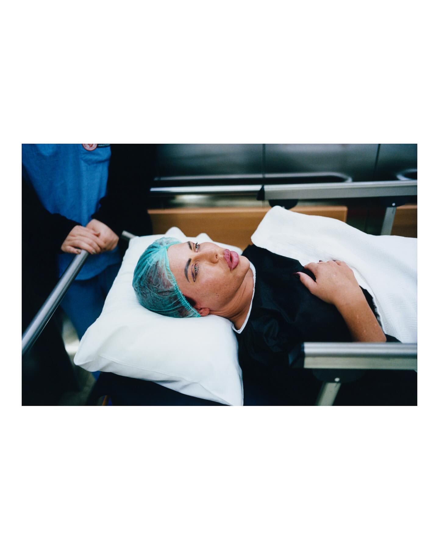Adult female patient lying on a hospital gurney with a surgical cap and pillow, being transported by staff member wearing scrubs.