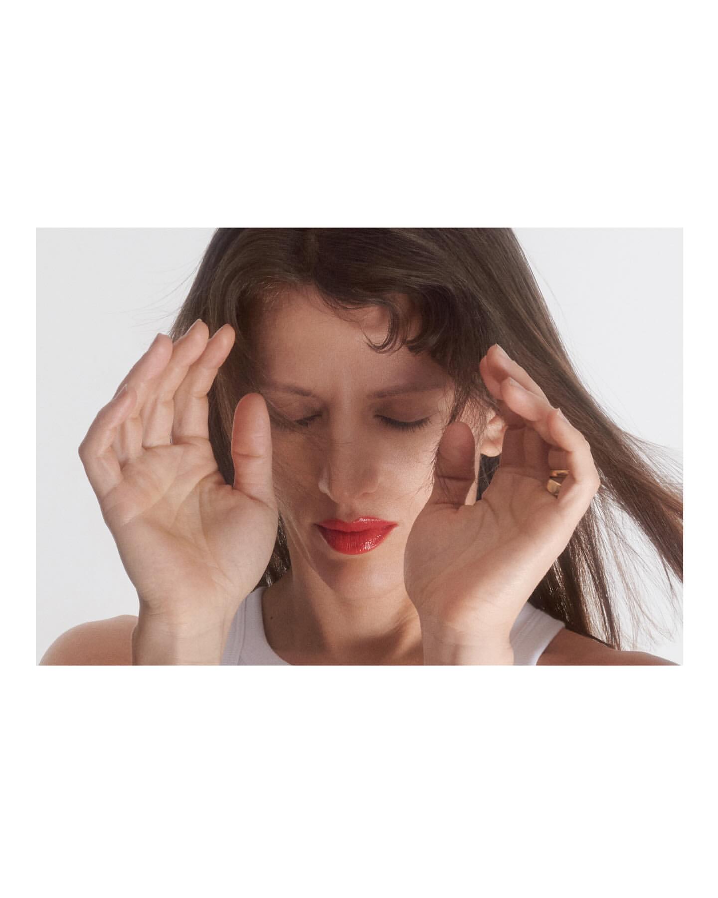 Close-up of a young woman with long brown hair and bright red lipstick, eyes closed, hands near her face, indoor studio setting, white background.