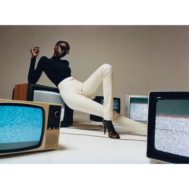 A dark-skinned model poses in a black top and cream pants, surrounded by retro TVs displaying static in a fashion editorial shot.