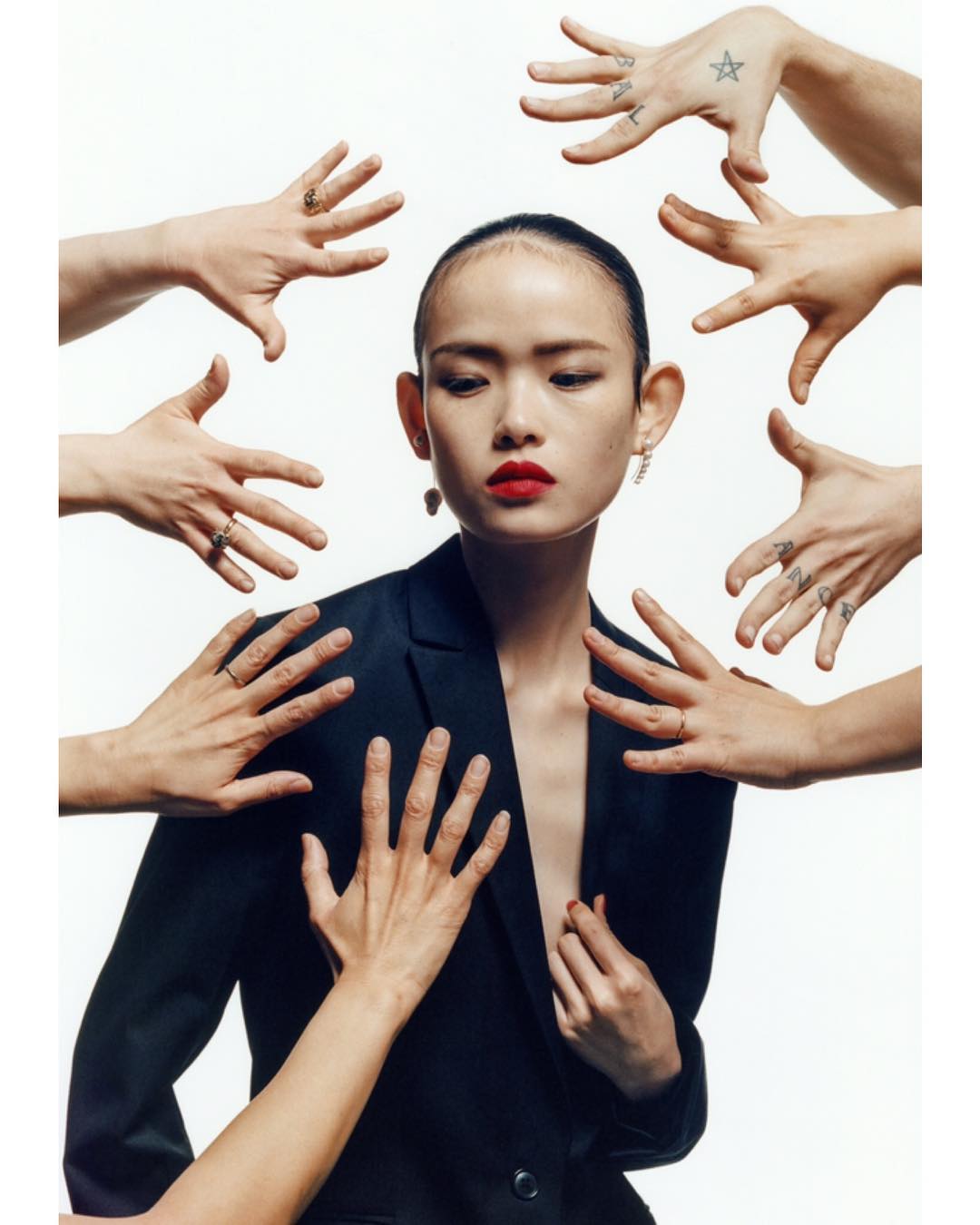 Portrait of an Asian woman in a blazer surrounded by reaching hands with tattoos in a brightly lit photo studio setting.