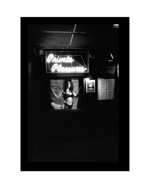 Monochrome image of a woman in lingerie visible through a window, under a 'Private Pleasures' neon sign in a dark, indoor setting.