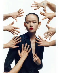 East Asian woman in a black blazer with red lipstick surrounded by many reaching hands against white background