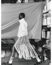 Black model wearing white jacket and fringed knit skirt walking past stacked fabric shelves in a monochrome editorial photo by Hanna Moon
