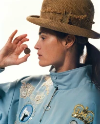 Profile portrait of a woman wearing a brown straw hat holding up a small coin, shot by Hanna Moon against a plain white background.