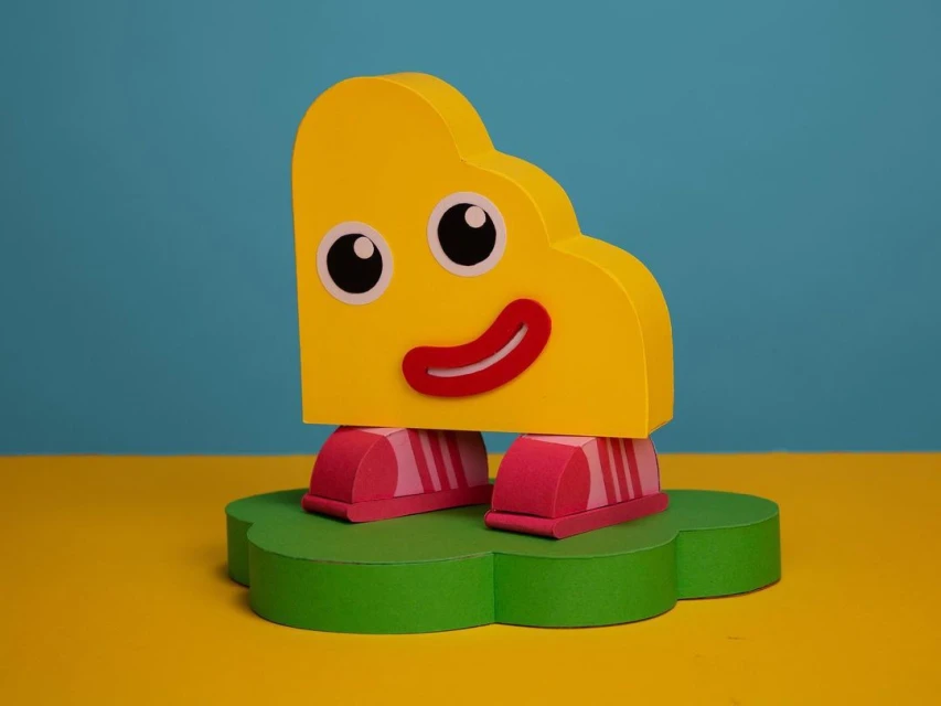 Cartoon-like figurine with a yellow block-shaped head, large eyes, and a red smile, stands on pink striped feet atop a green cloud base.