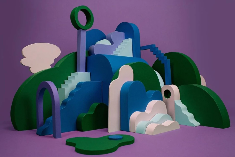 Abstract landscape installation with green hills, blue architectural shapes, staircases, and a lavender background, showcasing a creative design.