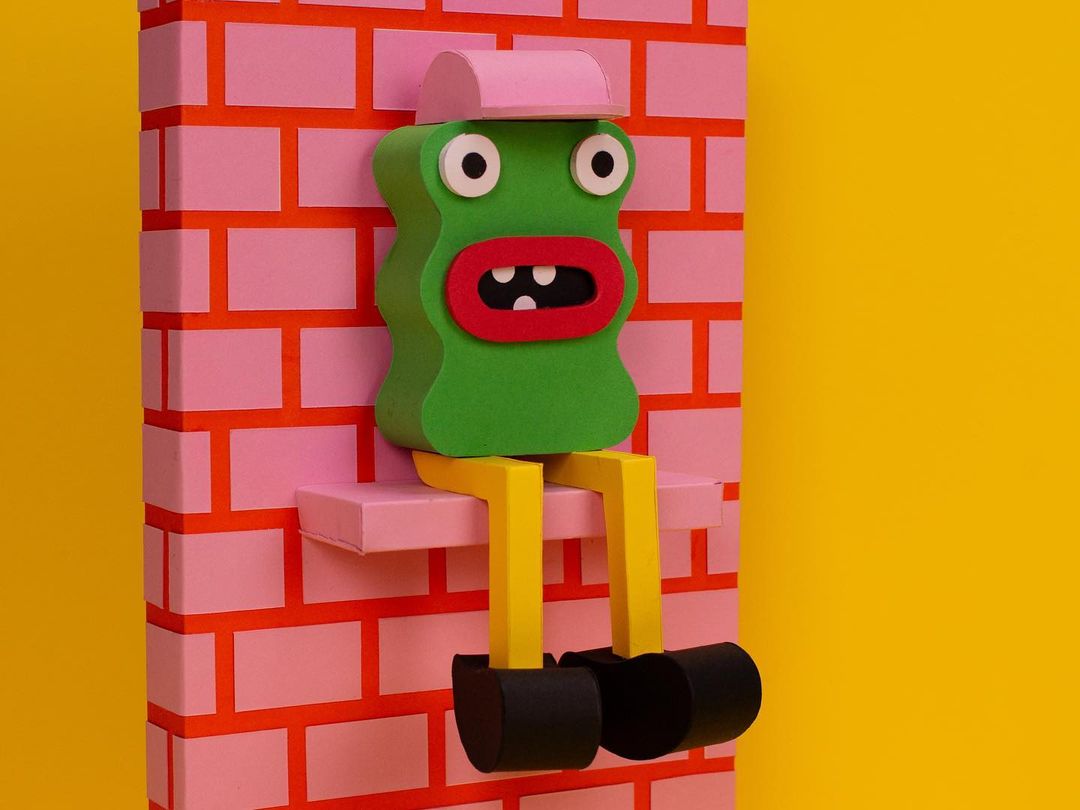 Whimsical, green, 3D paper art figure with a pink hat sits on a shelf against a pink brick wall and yellow backdrop in a playful scene.