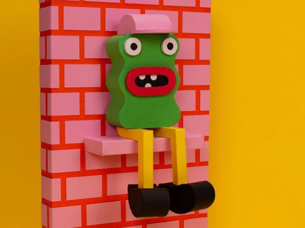 Whimsical, green, 3D paper art figure with a pink hat sits on a shelf against a pink brick wall and yellow backdrop in a playful scene.