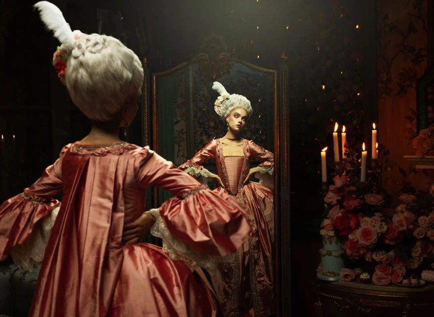 Woman with powdered wig and pink gown looks in ornate mirror, surrounded by candles and flowers in a dark, luxurious indoor setting.